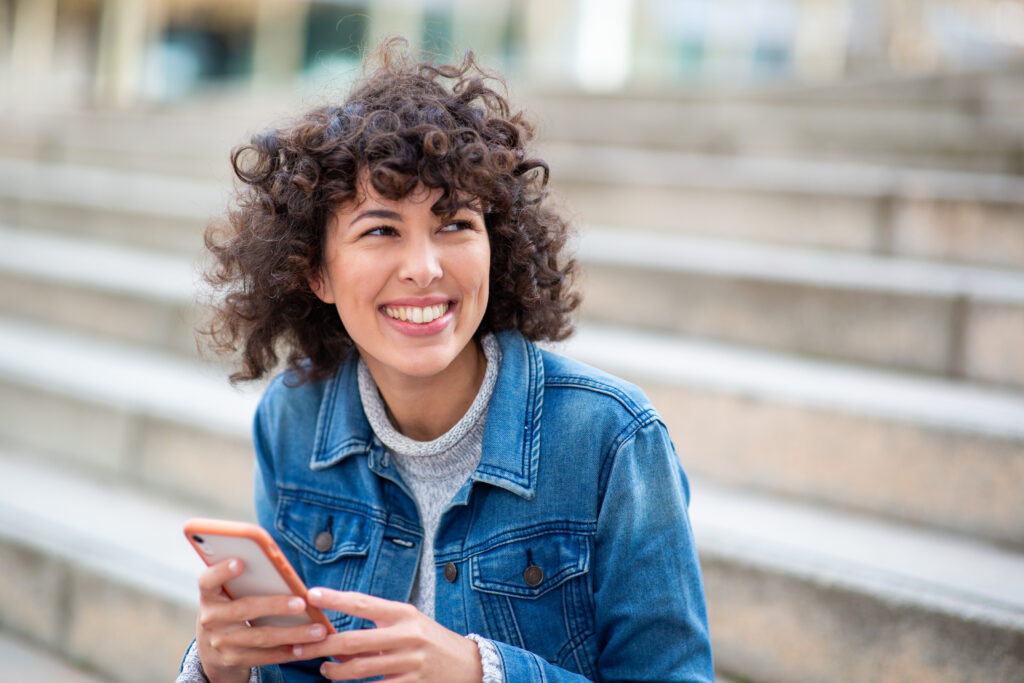 A young woman with curly hair wearing a denim jacket and holding a smartphone while smiling and sitting on outdoor stairs